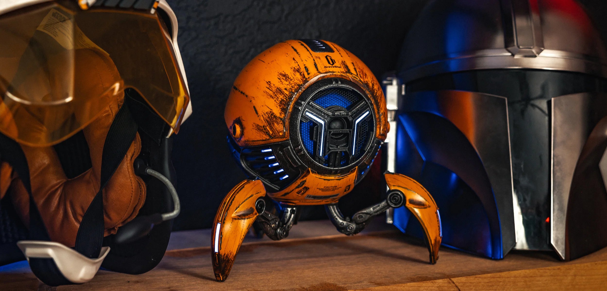 GravaStar Mars Pro Special Edition War-Damaged Yellow handcrafted Bluetooth speaker displayed with sci-fi helmets, featuring custom battle-worn finish, zinc alloy shell, 20-watt dual speaker system with passive bass radiator, and ice blue RGB lighting.