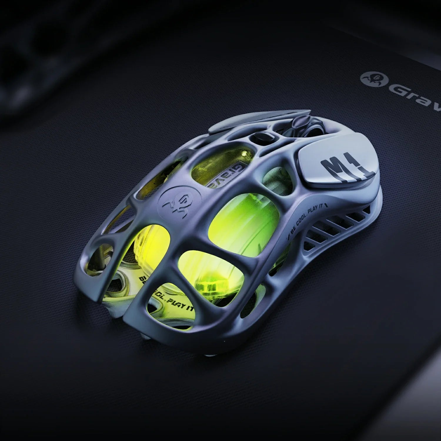 GravaStar Mercury M1 Pro Wireless Gaming Mouse Gunmetal Gray front-top perspective view featuring magnesium alloy skeletal frame, M1 logo on right button, BE COOL PLAY IT text, and green RGB backlighting.