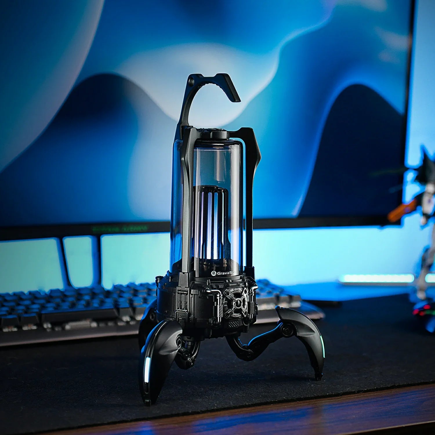 GravaStar Supernova Matt Black mecha lantern Bluetooth speaker on gaming desk featuring 25W dual-driver sound system, zinc alloy chassis, top carry handle, foldable tripod stand, and ice blue RGB lighting.