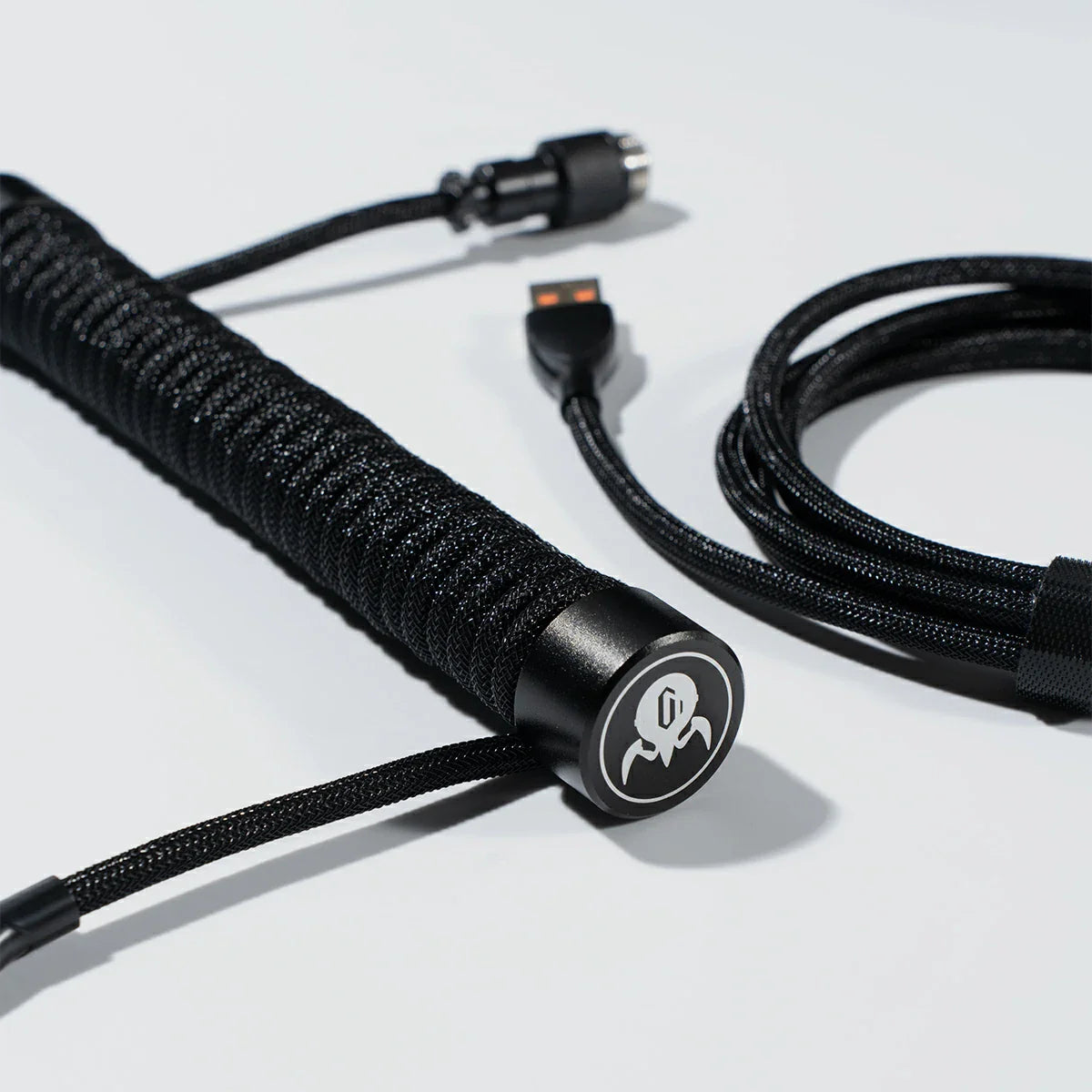 GravaStar Coiled Aviator USB Cable in black studio macro view showing GravaStar tactical logo emblem ring, 4-pin nickel-plated aviator connector, TPU EVA double-sleeved braided cable body, and gold-plated terminals, perfect paired with Mercury K1 Pro keyboard and GravaStar Gaming Mouse Pad deskmat.