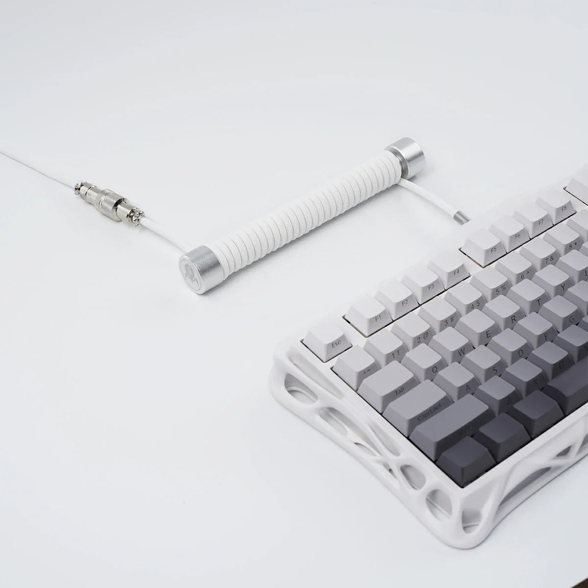 GravaStar Coiled Aviator USB Cable in white studio desktop top-down close-up view connected to Mercury K1 keyboard, featuring 4-pin nickel-plated aviator connector, TPU EVA double-sleeved braided cable body, and GravaStar tactical logo emblem ring, perfect paired with Mercury K1 Pro keyboard and GravaStar Gaming Mouse Pad.