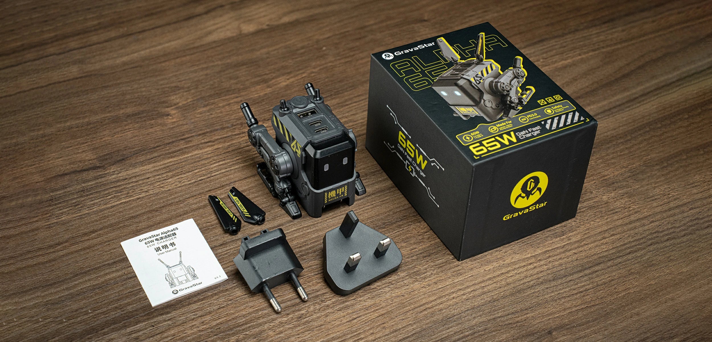 GravaStar Alpha65 GaN 65W Wall Charger Charcoal Gray packaging box studio view displaying matte black mecha robot body, dual antennas, UK and EU plug adapters, and user manual.
