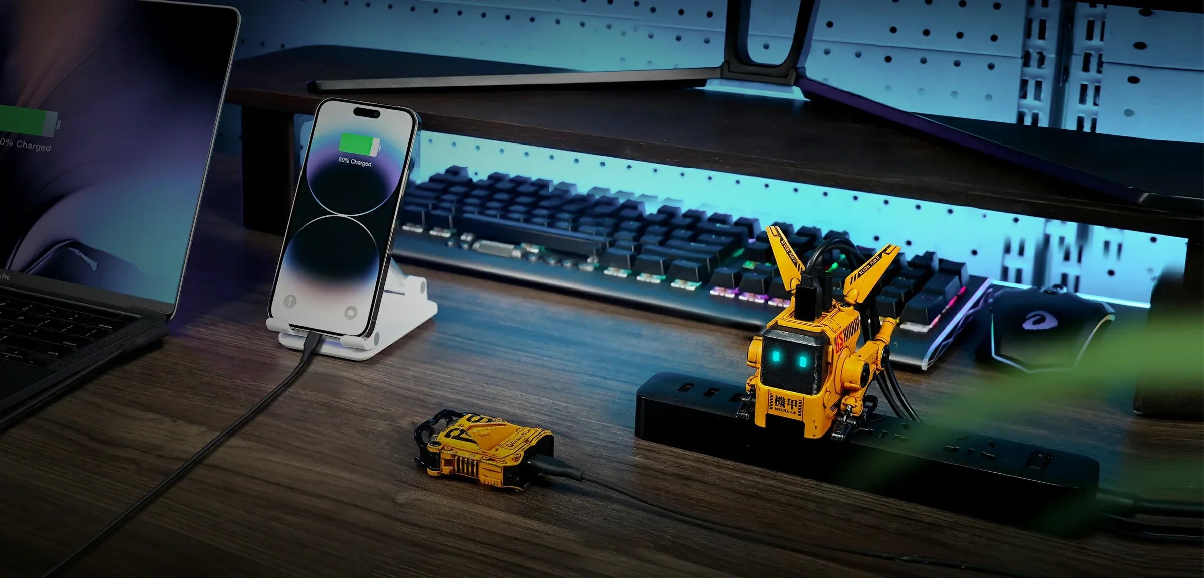 GravaStar Alpha65 GaN 65W Wall Charger War-Damaged Yellow cyberpunk desktop setup view charging smartphone alongside Sirius Plus earbuds case and Mercury K1 Pro gaming keyboard.