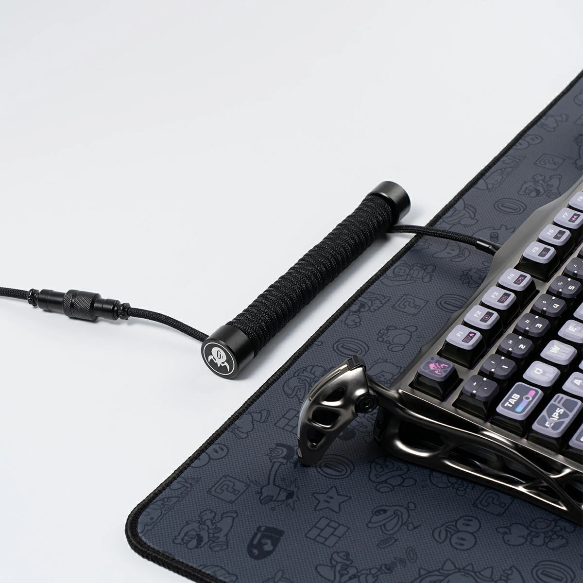GravaStar Coiled Aviator USB Cable Black studio setup view showing 4-pin GX12 aviator connector, TPU EVA double-sleeved braided cable body, GravaStar tactical logo emblem ring, connected to Mercury K1 Pro keyboard on gaming mouse pad deskmat.