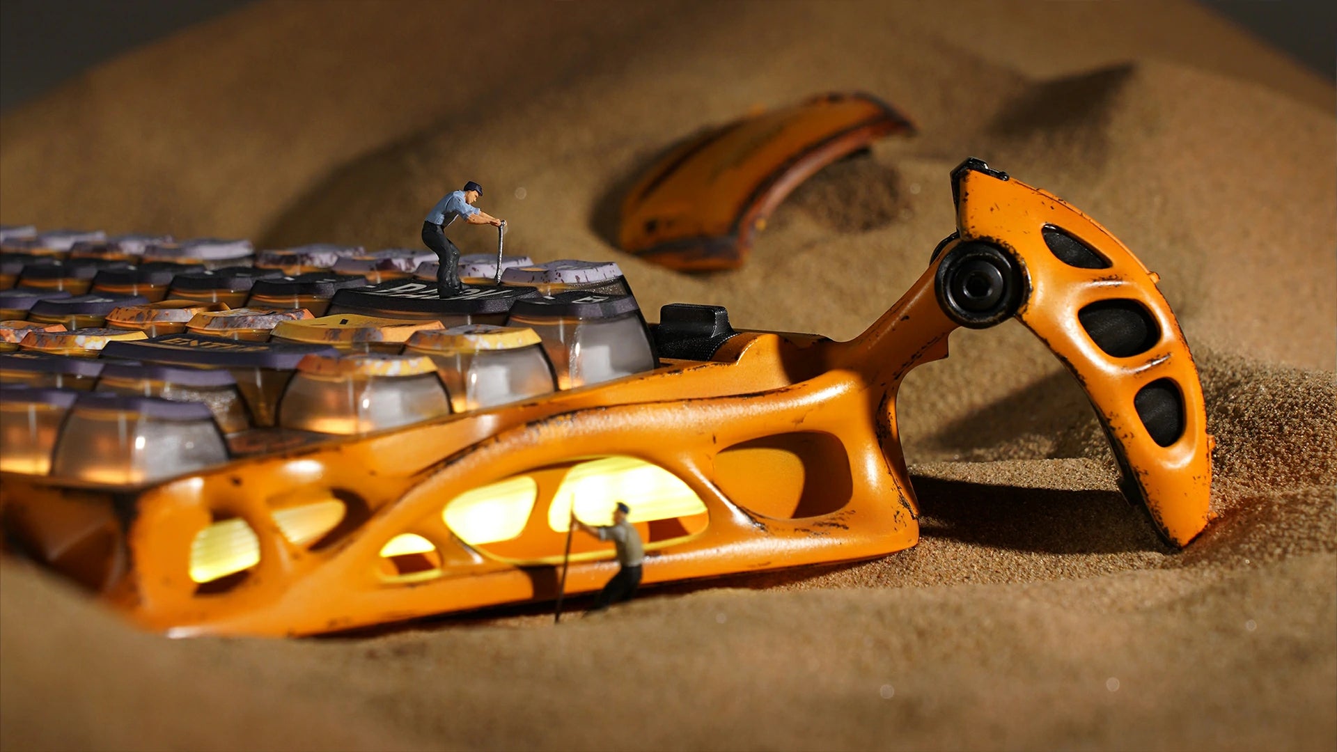 GravaStar Mercury K1 Pro Special Edition Battle-Worn Yellow 75% wireless mechanical gaming keyboard macro view featuring handcrafted weathered aluminum exoskeleton embedded in sand dune with miniature worker figures and warm RGB lighting.