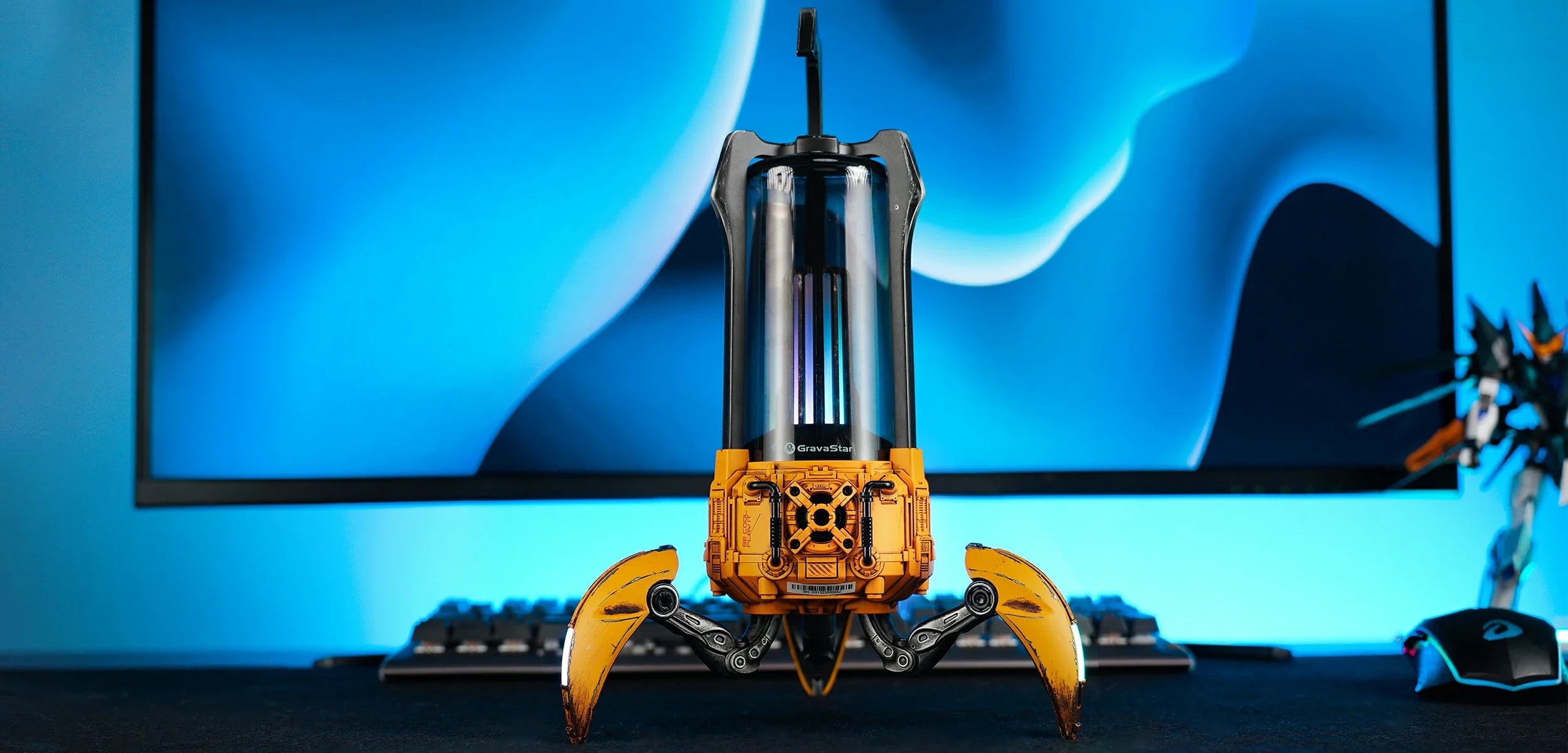 GravaStar Supernova War Damaged Yellow handcrafted mecha lantern Bluetooth speaker full width setup banner featuring 25W dual-driver sound system, zinc alloy frame, 3-legged tripod stand, top carry handle, and ice blue RGB lighting alongside gaming keyboard and mouse.