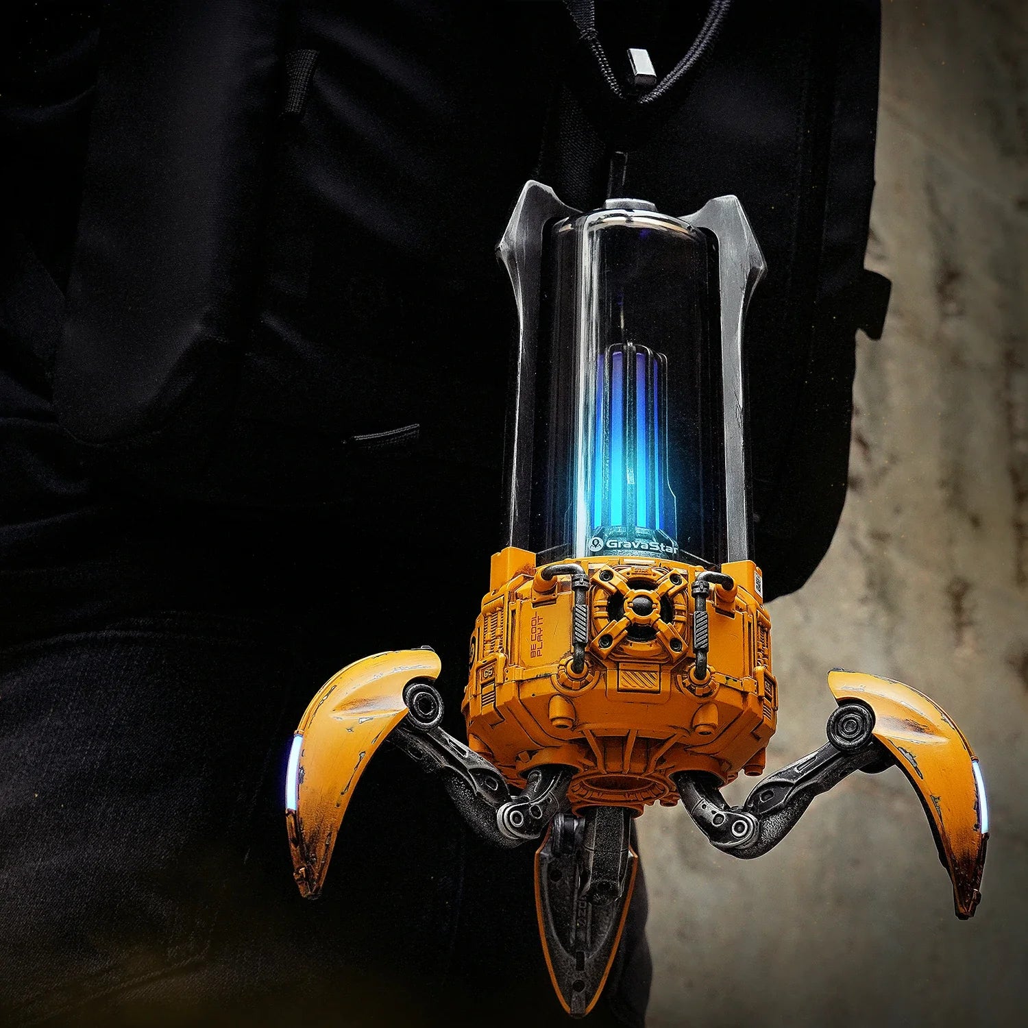 GravaStar Supernova War Damaged Yellow mecha lantern Bluetooth speaker attached to tactical backpack featuring removable carry handle, lightweight 1.6kg frame, 25W dual-driver sound system, foldable tripod stand, and ice blue RGB lighting.