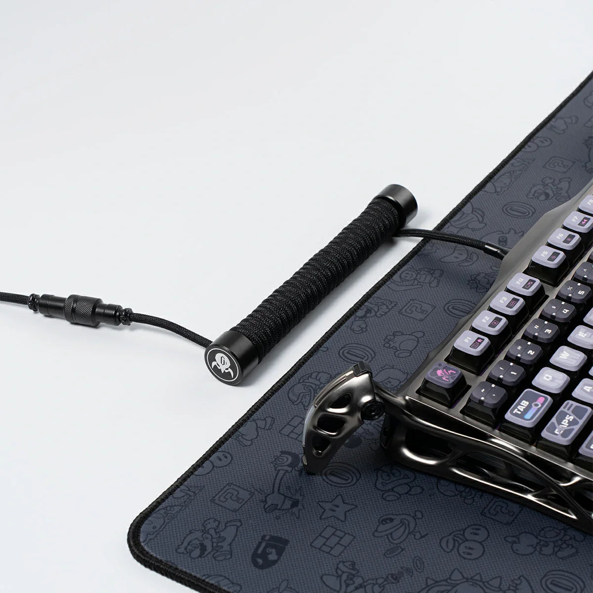 GravaStar Coiled Aviator USB Cable in black studio desktop setup side-angle perspective view connected to Mercury K1 Pro keyboard, featuring 4-pin nickel-plated aviator connector, TPU EVA double-sleeved braided cable body, and GravaStar tactical logo emblem ring, perfect paired with Mercury K1 Pro keyboard and GravaStar Gaming Mouse Pad.