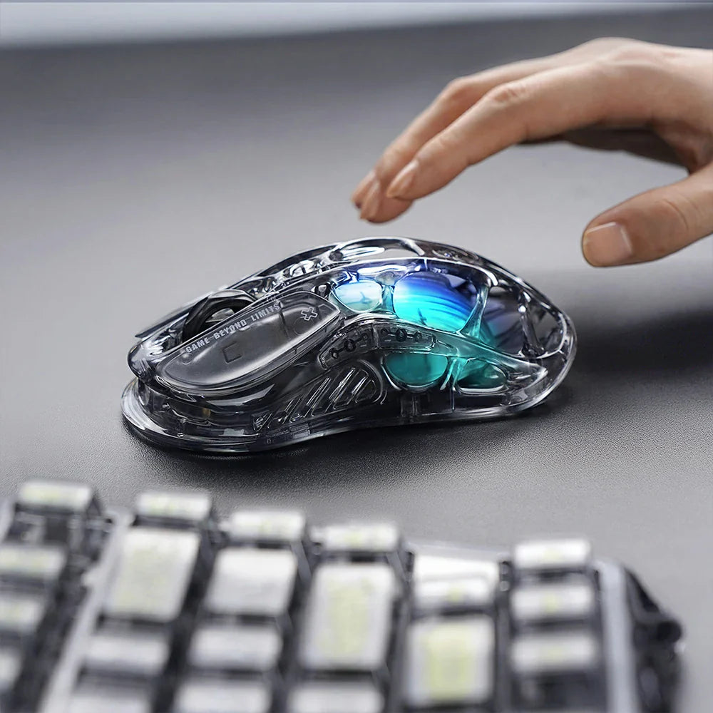 GravaStar Mercury M2 Transparent Black wireless gaming mouse in-hand ergonomic grip view on desk setup with Mercury K1 Lite keyboard – Highlighting see-through crystal housing, PAW3395 26K DPI sensor, Kailh 80M micro-switches, 2024 Red Dot Award design, and blue RGB lighting.
